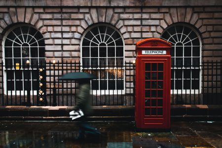 time lapse photography of woman walking on street while holding umbrella near London telephone booth beside wall
